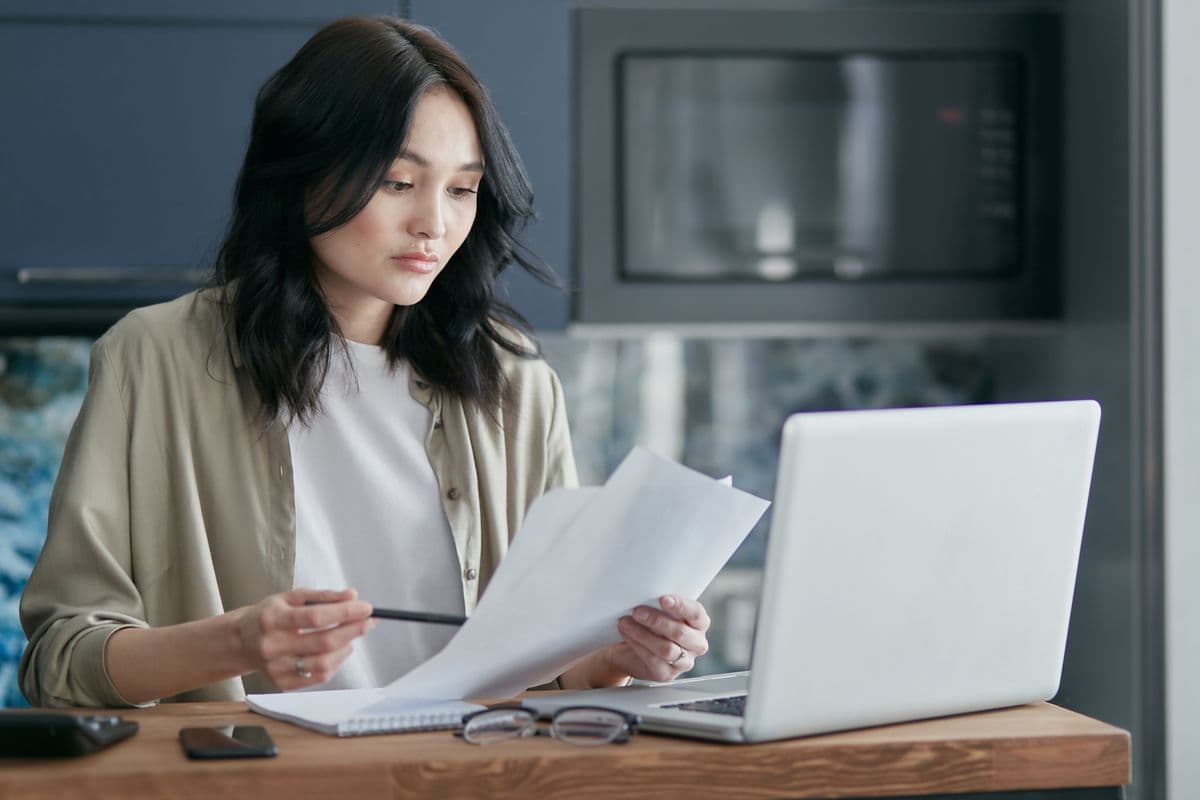 Mujer revisando un documento en la barra de la cocina, con laptop y celular