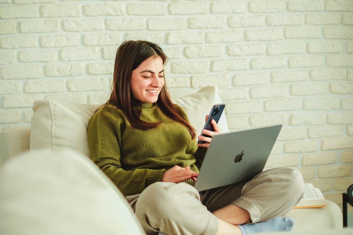 Mujer sentada en el sofa con la laptop sobre las piernas, comparando algo en la pantalla del celular