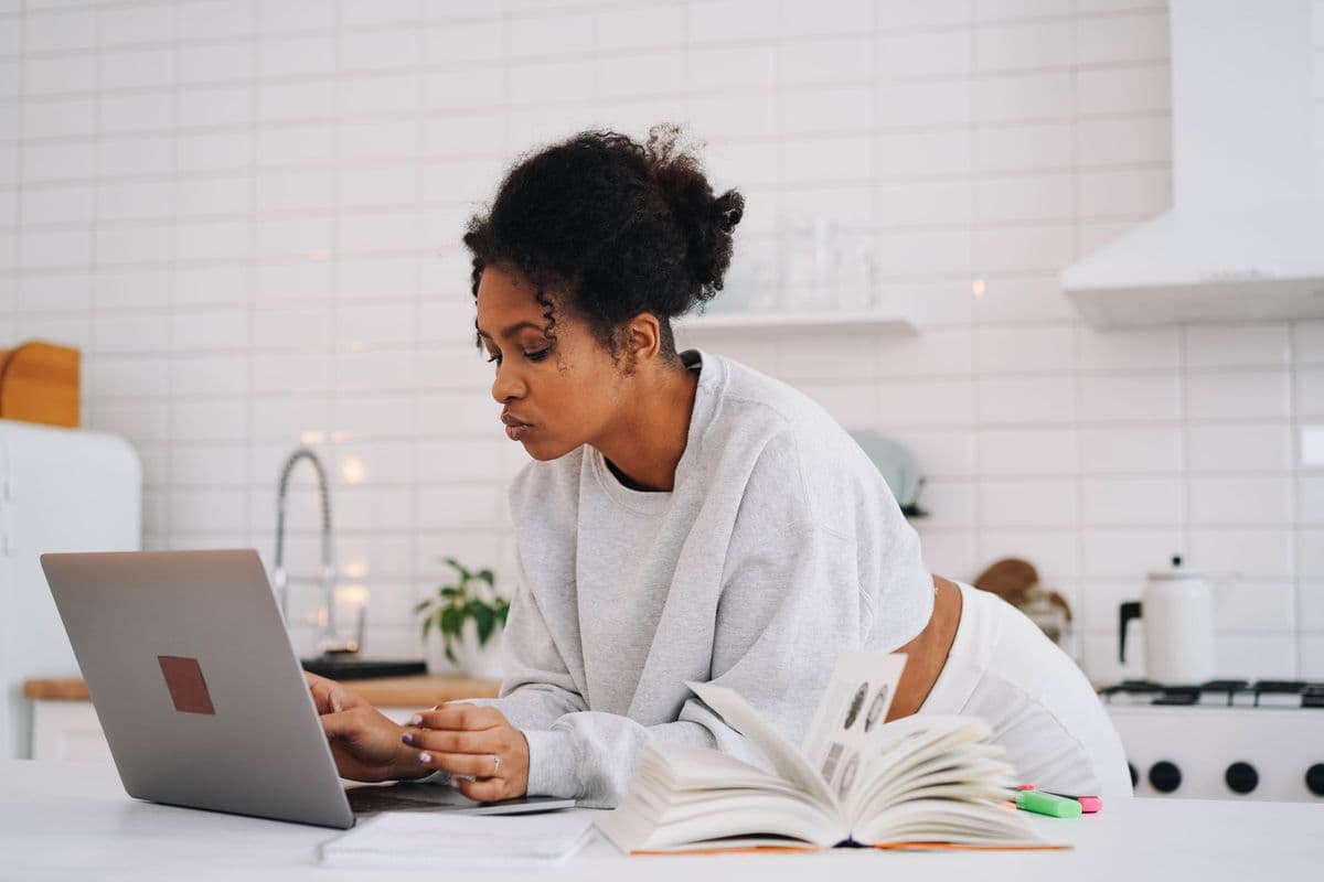 Mujer apoyada en la barra de la cocina leyendo su laptop, con un libro de consulta y una libreta al lado