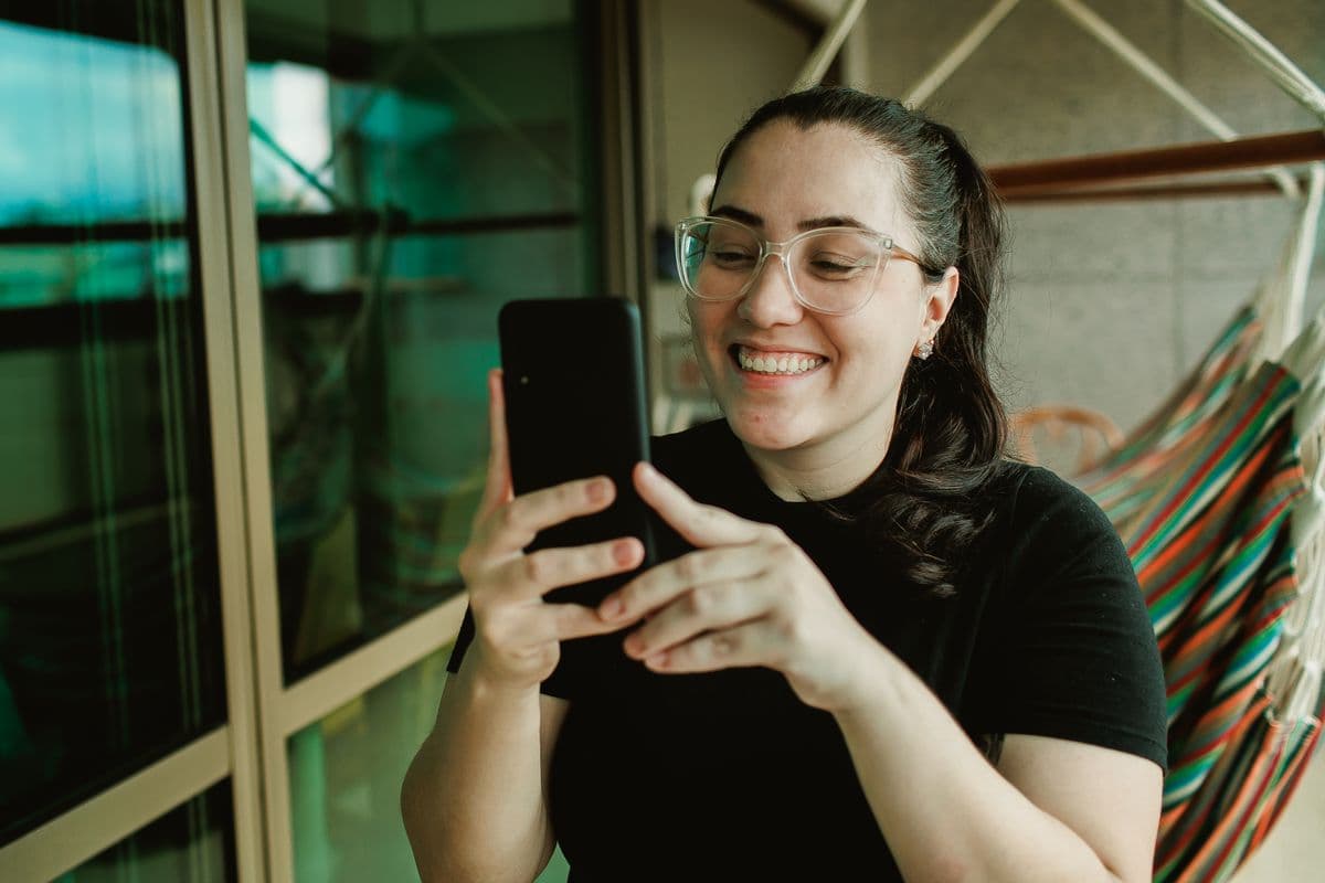 Mujer con lentes sentada en una silla colgante en el balcon, riendo de lo que ve en su celular