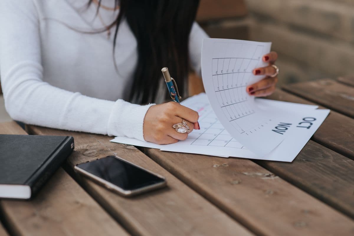 Manos llenando un calendario de papel sobre una mesa de madera, con celular y libreta al lado